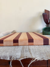 Load image into Gallery viewer, A striped wooden cutting board made of alternating light and dark wood tones highlighting the edge joinery, resting on a burlap-covered wooden surface in an indoor setting.