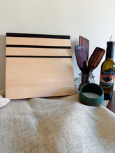 Load image into Gallery viewer, A square wooden cutting board with three horizontal dark stripes, standing upright on a beige linen cloth next to kitchen utensils and a bottle of olive oil.