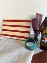 Load image into Gallery viewer, A striped wooden cutting board with alternating light and dark wood bands, displayed on a beige linen runner next to kitchen utensils, a small teal bowl, and a bottle of olive oil.
