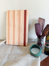 Load image into Gallery viewer, A striped wooden cutting board with alternating light and dark wood grains stands upright on a beige linen table runner. To the right are kitchen props including a glass jar with wooden spoons, a small teal bowl of salt, and a bottle of olive oil against a plain off-white wall.
