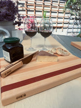 Load image into Gallery viewer, A wooden charcuterie board with alternating light and dark wood stripes, styled with a slice of cheese, crackers, a jar of preserves, and a wooden knife. Two glasses of red wine and a patio background are visible in the soft-focus setting.
