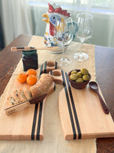 Load image into Gallery viewer, The paired wooden serving boards with black stripes, styled with bread, sliced bread, oranges, olives, a spoon, and a small jar on a table runner in a bright indoor setting.
