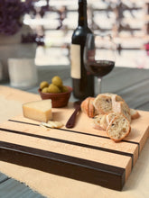 Load image into Gallery viewer, A wooden serving board with dark brown end caps and dark horizontal stripes, styled with sliced bread, a wedge of cheese, a small bowl of olives, a wine bottle, and a glass of red wine on a table.
