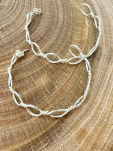 Load image into Gallery viewer, A pair of silver wire hoop earrings with a twisted, knotted design, displayed on a textured wood slice background.
