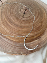 Load image into Gallery viewer, A silver crescent moon pendant necklace on a delicate silver chain, displayed on a rustic wooden slice coaster over a light fabric background.
